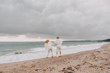 Women sea walk friendship spring. Two girlfriends, redhead and blonde, middle-aged walk along the sandy beach of the sea, dressed in white clothes. Against the backdrop of a cloudy sky and the winter