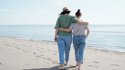 Young romantic lesbian couple walking on sea beach barefoot embracing each other by the waist