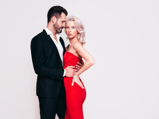 Beautiful fashion woman and her handsome boyfriend in black elegant suit. Sexy blond model in red evening dress. Fashionable couple posing in studio. Brutal man and his female. Isolated on white