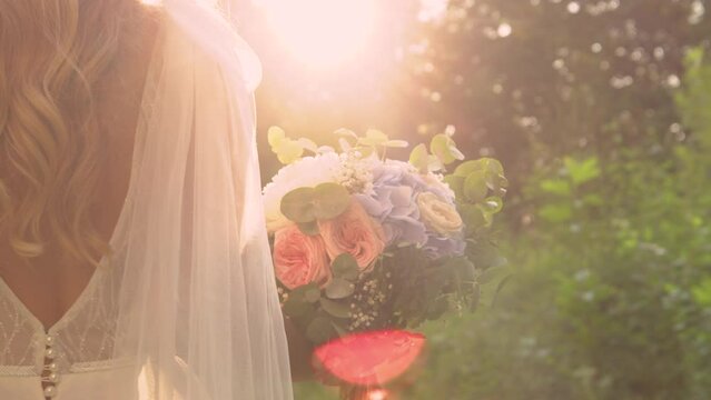 LENS FLARE, CLOSE UP Golden light and bride walking with gorgeous bridal bouquet. Magnificent wedding bouquet with summer flowers in pastel shades and beautiful bride in white dress at golden sunset.