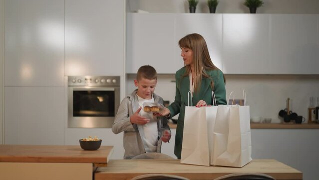 Mother and son unpack shopping bags with food standing in kitchen. Hungry schoolboy takes muffins and waffles looking at food with happy expression