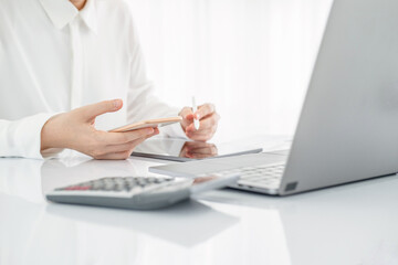 Asian businesswoman in white formal clothes writing note in tablet and checking smart phone. Business woman busy working with documents and tablet  in workstation. close up clean image.