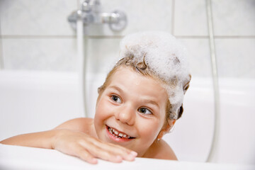 Cute child with shampoo foam and bubbles on hair taking bath. Portrait of happy smiling preschool girl health care and hygiene concept. Washes hair by herself.