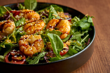 Salad with greens, tomatoes and grilled shrimp in a black plate. Wooden background
