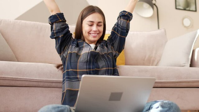 Young woman sits on floor in lotus position with laptop, looks at screen and smiles admiringly, happily raises hands to top and laughs. Get good news, emotions. Receipt salary, good exam results.