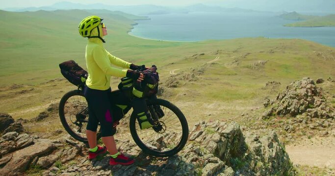 The woman beside her bike in the mountains, rests from her cycling journey. Girl looking at the stunning view of lake by mountains. Concept touring bicycle bikepacking bags, travel cycling adventure
