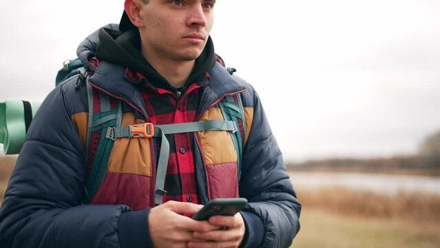 Portrait of young guy tourist backpacker in cap with smartphone finds route on map hiking in autumn. He looks around in search of right path. Hike, tourism, travel, explore wild nature concept.
