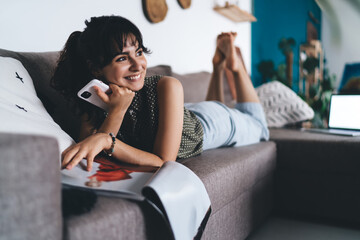 Cheerful woman with smartphone and magazine relaxing on sofa