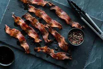 Wavy strips of crispy maple cracked peppercorn bacon on a black slate board. - obrazy, fototapety, plakaty