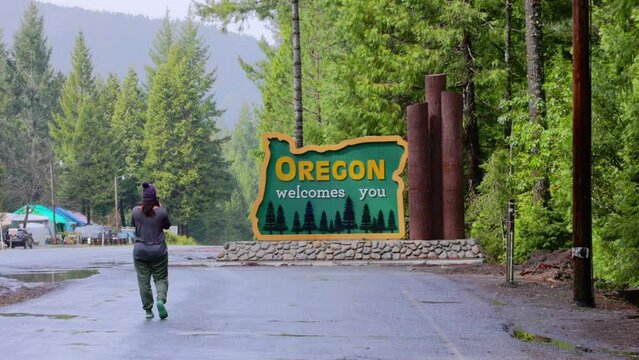 Woman, 30s, walks up and takes photo of Oregon welcomes you sign on Redwood Highway.