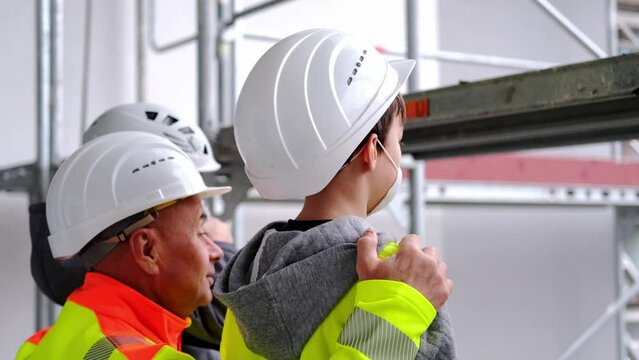 experienced male builder in yellow protective jacket, children in white hardhats came on excursion, father, grandfather tells grandchildren about his profession with background of high buildings