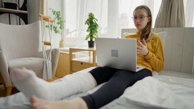Teenage girl with broken leg in cast sits at home and use laptop to online video call conversation with friends. Young woman sitting on bed in living room next to crutches. Injury, home treatment.
