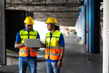 Two men warehouse indian worker wearing safety hardhats helmet working on laptop computer in warehouse of Wholesale Merchandise. shelves, pallets and boxes. Container and warehouse factory