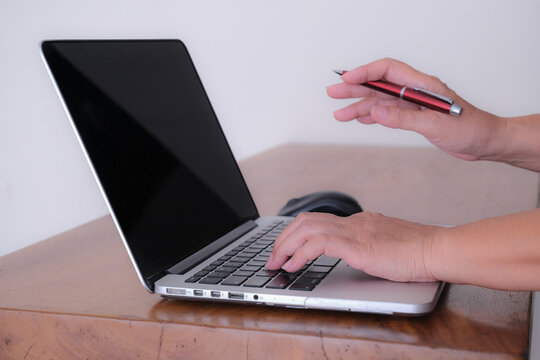 Close up, Side view: A woman's hand on laptop keyboard, other hand holding a pen.