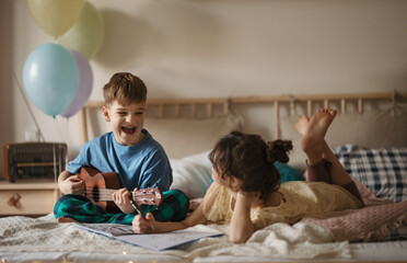 Little boy plaing on the guitar, while his sister drawing a picture.