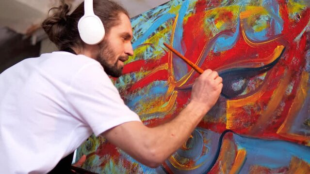 An imaginative artist, clad in a white t-shirt and black apron, donning headphones, is crafting an abstract painting on a large canvas with a vibrant palette while reveling in his favorite music.