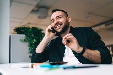 Cheerful man sitting at wooden counter and talking on smartphone in office