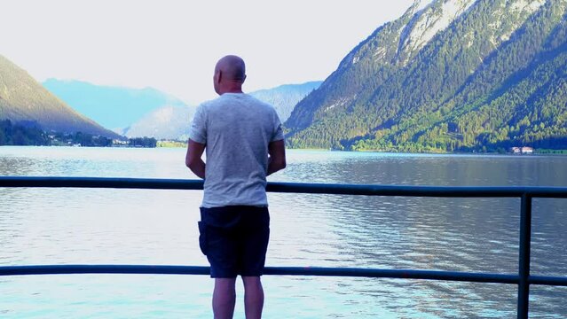 mature man 60 years old stands on observation deck, admires mountain landscape, lake Achensee in Austria, green mountains rises above calm expanse of water, vacation by reservoir, resort place tyrol