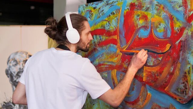 Innovative talented artist with long hair in headphones, wearing a black apron smeared with paint, creates masterpiece using red blue yellow paints, painting large canvas in creative studio.