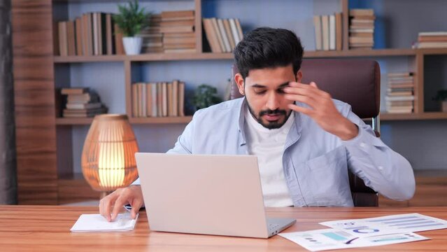 Overstressed, humorous Indian businessman rushes around his office desk, struggling to meet a deadline. He picks up documents, checks his laptop, writes notes, hurries to complete his tasks on time.
