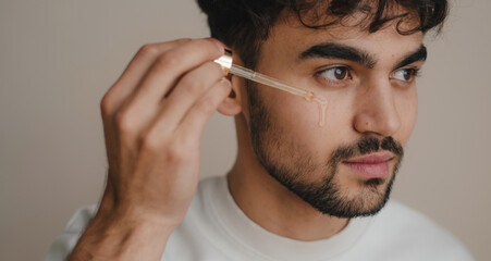 Attractive young man applying essential oil on facial skin while looking away isolated on plain...