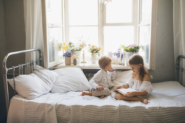 Cute boy and girl sitting on bed reading book together near window.