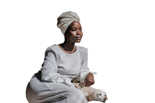 Peaceful African young woman in white turban and dress sitting against transparent background strokes white dog. Friendship of owners and pets. Pensive 
