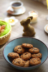 Easter eggs in the basket, bowl of cookies, chocolate pralines, Easter bunny figurine, cups of tea, glasses of juice, flowers and lit candles on the table. Selective focus.