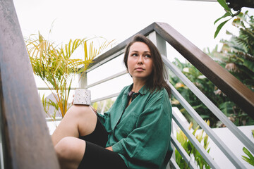 Pondering woman 30 years old with coffee cup resting outdoors at stairs, contemplative female dressed in casual green shirt thinking about cappuccino taste during daytime spending on weekend