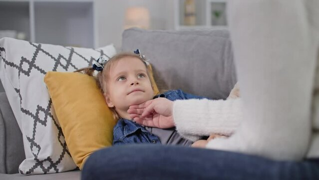 child health, little girl lies on a pillow and opens mouth showing throat to her mother lying on sofa in room