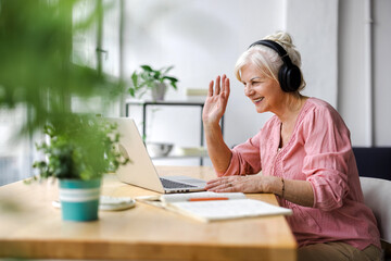 Happy senior woman in headphones using laptop and waving at camera during video chat
