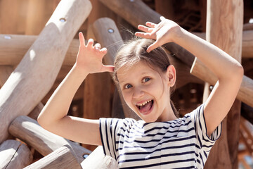 Grimacing Child making Grimaces with Ten Fingers as Horns Having fun Playing Outdoor on Wooden Playground.