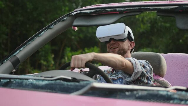 Man in virtual reality glasses driving wrecked car. Male gamer playing VR game with headset sitting in pink convertible outdoors , trees in background
