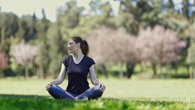 Doing yoga on nature - an adult woman in glasses sits in the lotus position and warming up her neck