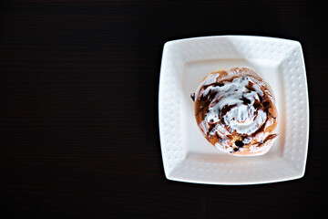 Cinnabon bun lies on a white square plate on a dark background. View from above. There is a place for text. Home cooking. Up close