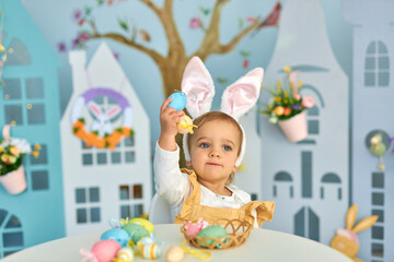 Baby girl celebrate Easter. Funny happy kid in bunny ears Sits by the table and playing with Easter eggs. Colorful Easter eggs and flowers. Home decoration and flowers