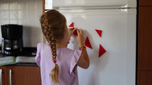Little blonde girl with long braid is playing with magnets on fridge, back view, kid playing with tangram