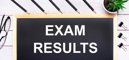On a light wooden table - a board with the text EXAM RESULTS, pencils, plants, glasses and paper clips. Business concept