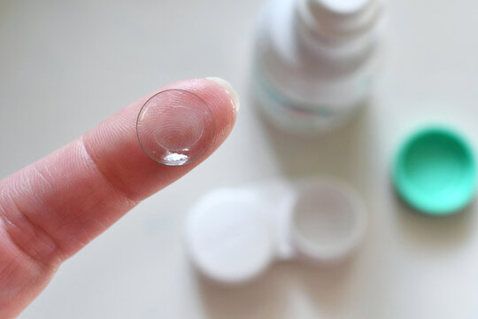 Person Holding Contact Lens On Finger With A Case And Solution Bottle In The Background. Top View