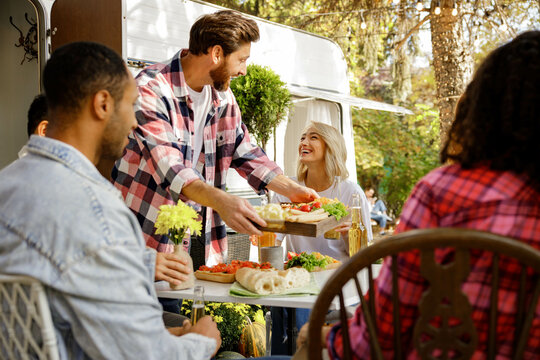A cheerful multinational company is having fun on a picnic. Multisex friends sitting at the table outdoors celebrating eating and drinking drinks.