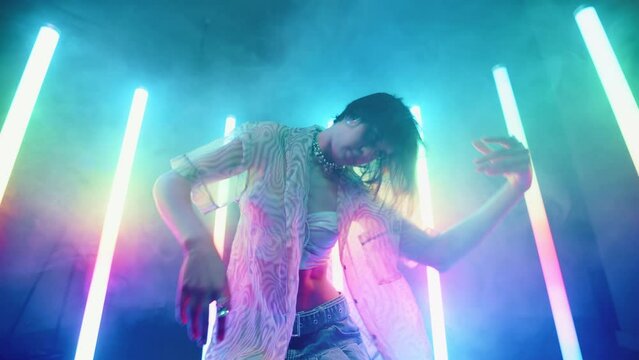 Stylish young woman dance with her hands backlit with colorful neon light lamps, female in trendy outfit dancing in night club, creative performer, haze and glowing tube lights, slow motion wide shot