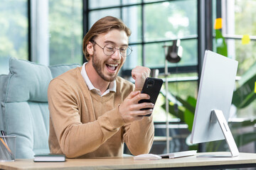 Happy young student in glasses sitting in office at home in front of computer, holding phone in hand, showing victory success gesture with hand, celebrating, smiling.