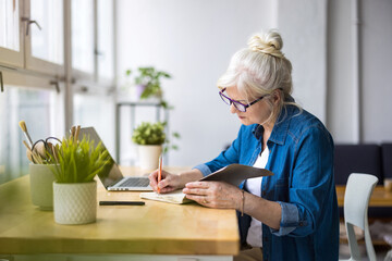 Smiling mature businesswoman writing in notebook while sitting at table in office