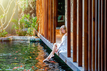Woman relax in luxury spa with fish pond, enjoy bright morning and nature, relaxing and having good time sitting on wooden bridge barefoot. female playing feet in water at outdoor pond. Slow motion