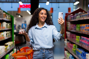 A young Latin American woman, blogger stands in a supermarket with a basket among the shelves with...