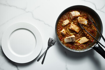 Frying pan with Indian salmon curry dish. Fried salmon fillets, stewed in curry sauce based on red tomatoes and other vegetables. Empty clean white plates and forks nearby. Top view. 