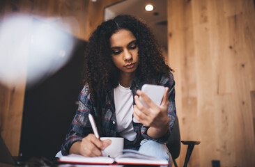 Fototapeta na wymiar Focused black woman taking notes while sitting with smartphone
