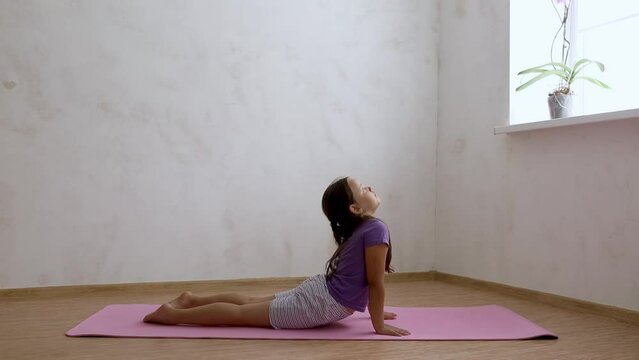 School age girl in sportswear bending back lying on floor with raised head on exercise mat in empty room, free copy space. Home workout, gymnastics, kids sport, stretching, activity.