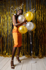 Smiling afro american woman in formal dress posing on gold decorations background in festive hall...