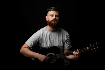 Portraite of handsome brunette bearded man musician, guitarist standing and holding a acoustic guitar in a hand and looks in a camera on a black background studio. Ready to play a music.